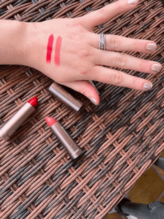 A hand with two red lipstick swatches on the back. Two open lipstick tubes are placed on a wicker surface beside the hand.