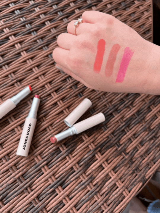 A hand with lipstick swatches in shades of pink, rose, and red is shown over a woven surface, with two opened lipstick tubes and a closed tube labeled "JONES ROAD" placed nearby.