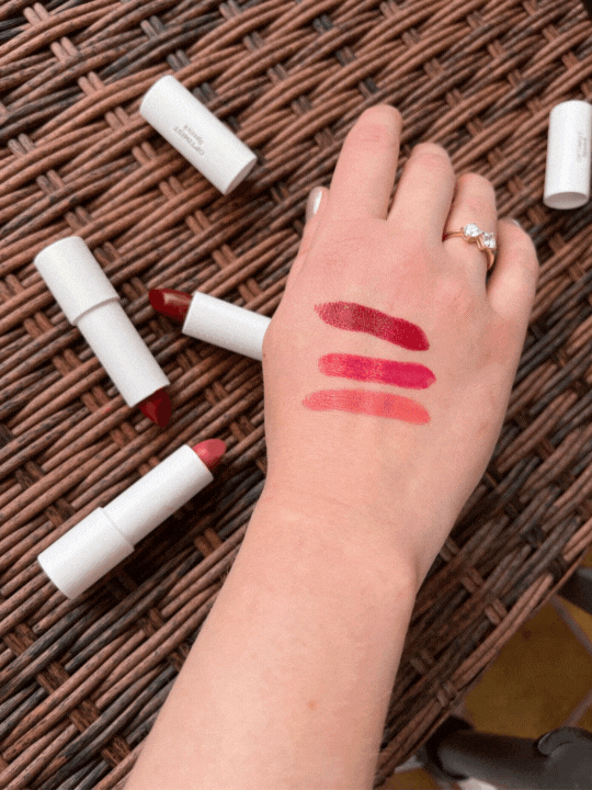 A hand with three swatches of lipstick, next to several opened lipsticks on a woven surface.