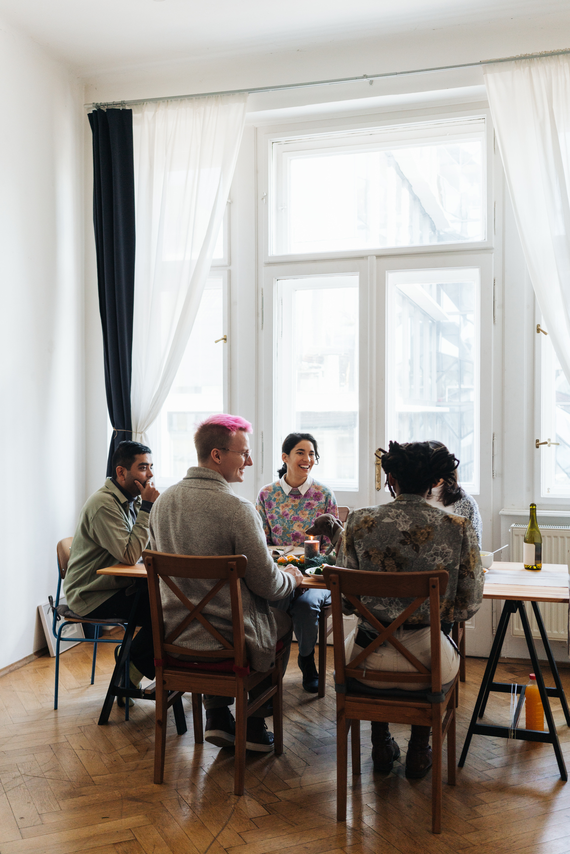 A group of five people sit around a table near a large window in a bright room, engaged in conversation.
