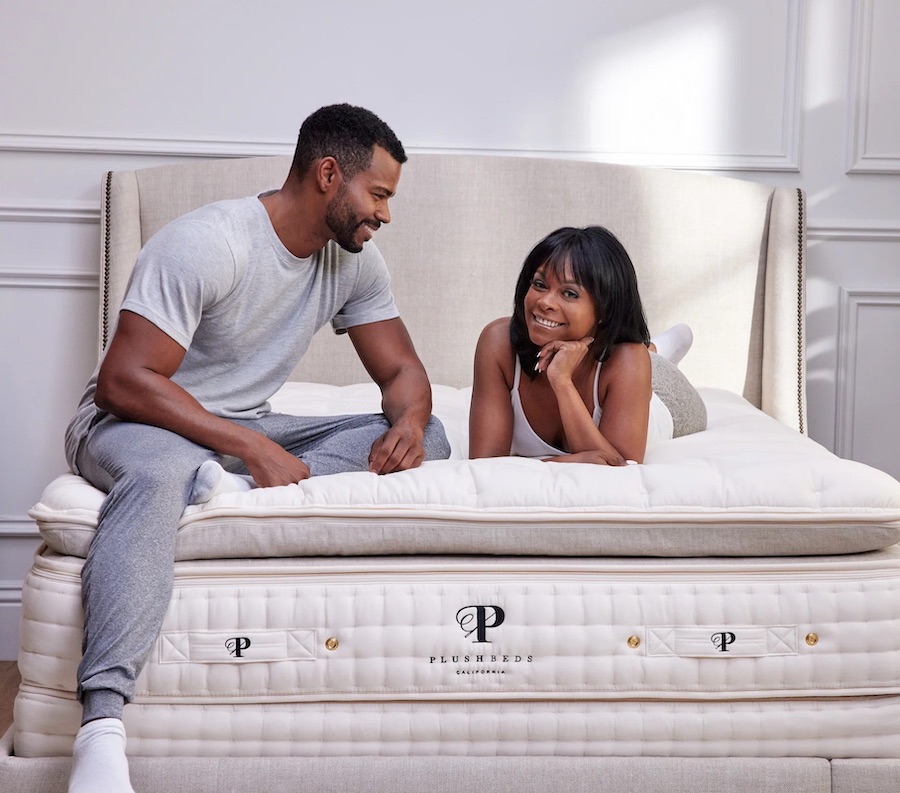 A man and woman smile while sitting and lying on an organic Plushbeds mattress in a bright, stylish bedroom.