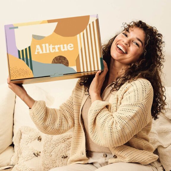 Woman sitting on a couch, smiling and holding a colorful Alltrue subscription box.