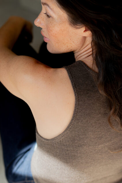A woman with freckles and long brown hair, wearing a sleeveless brown top and dark pants, sits turned slightly away from the camera.