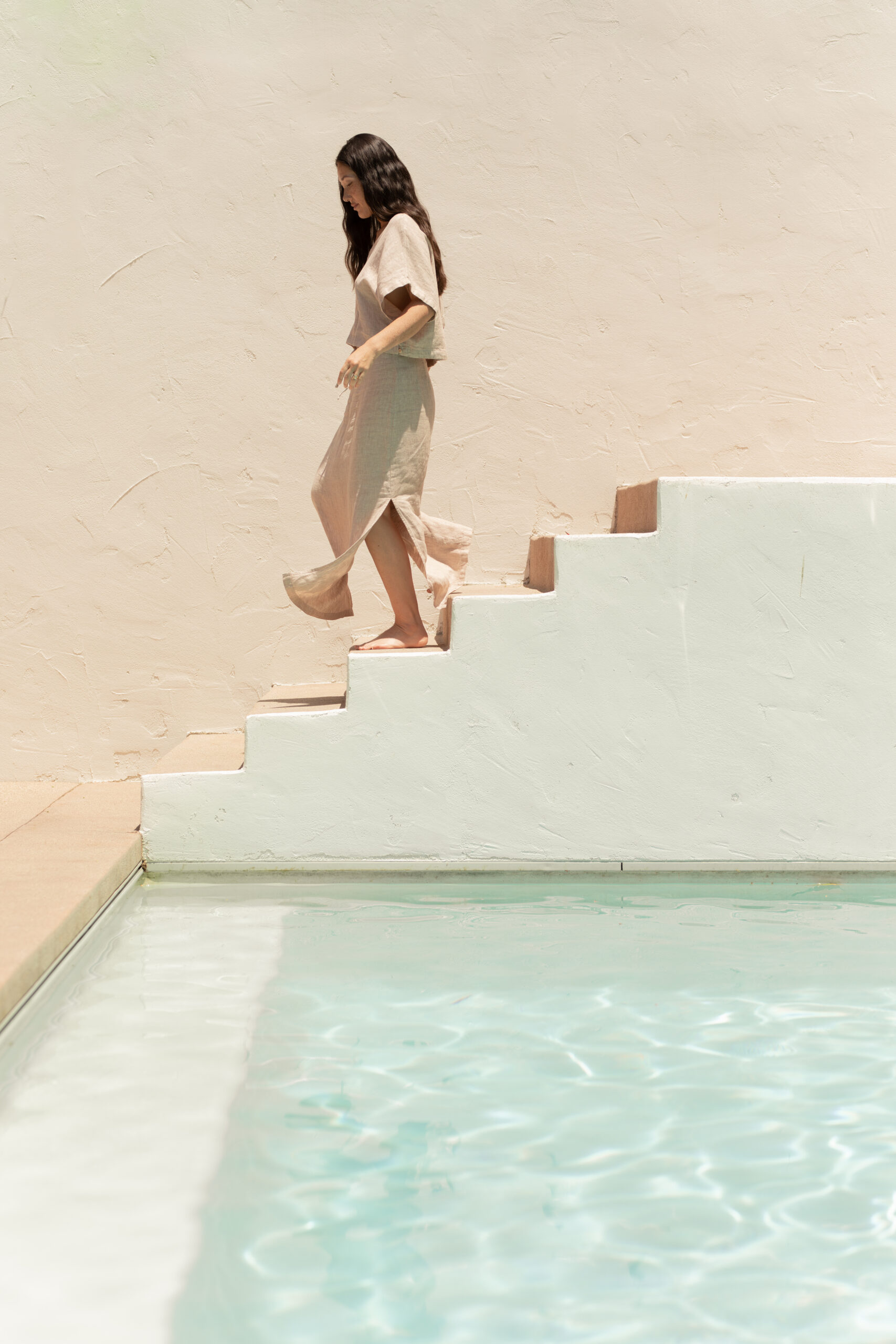 A woman in a light dress walks down white stairs beside a pool with clear water, against a beige textured wall.