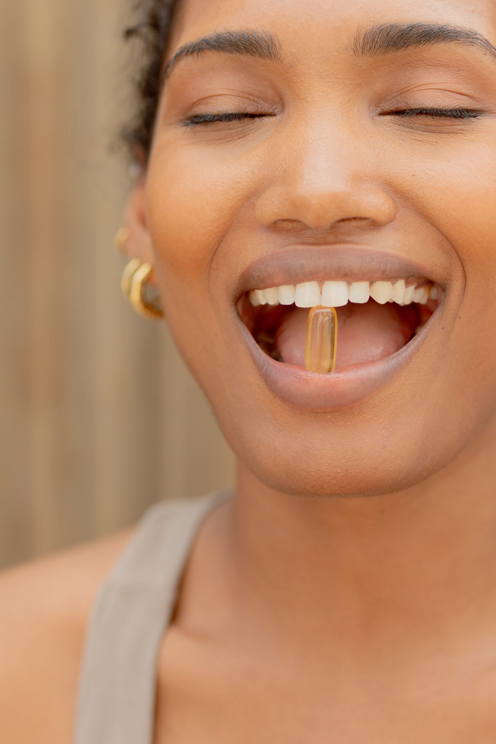 A person with closed eyes smiles while holding a yellow capsule between their teeth.