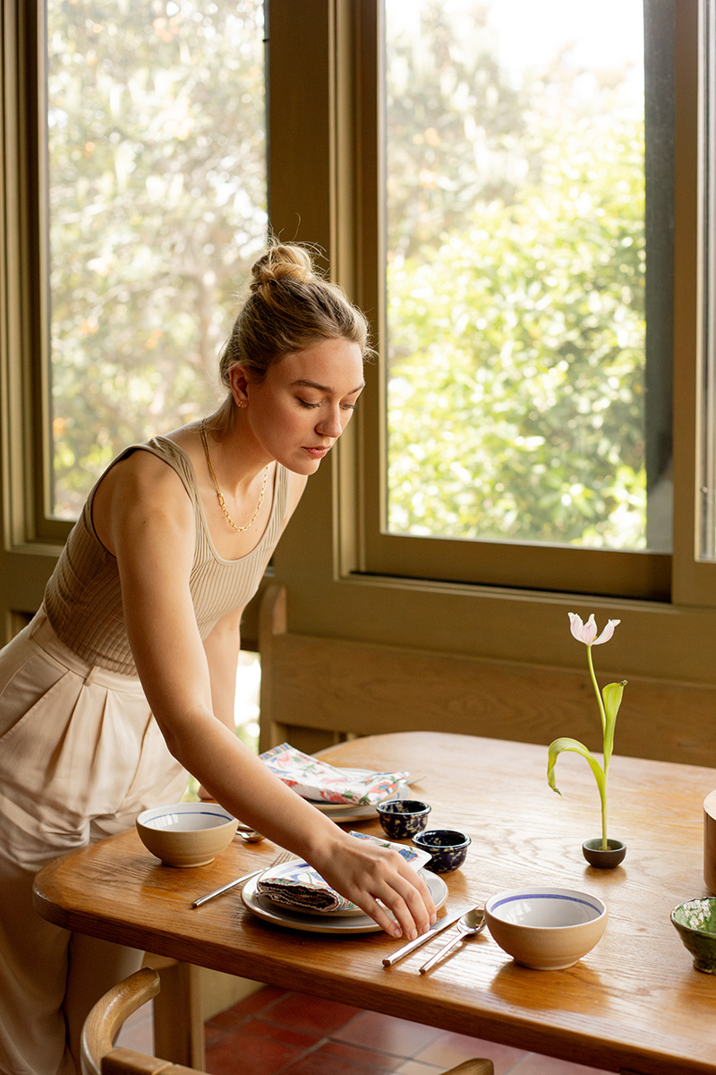 A person sets a table by a window with a view of greenery. The table has bowls, a plate, chopsticks, and a single tulip in a vase.