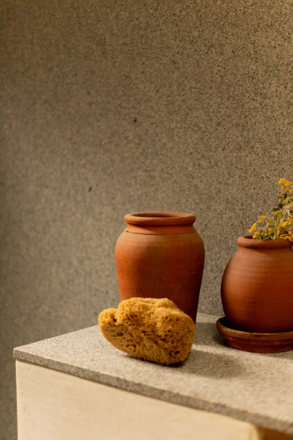 Two clay pots and a natural sponge rest on a gray countertop against a textured gray wall; one pot holds dried yellow flowers.