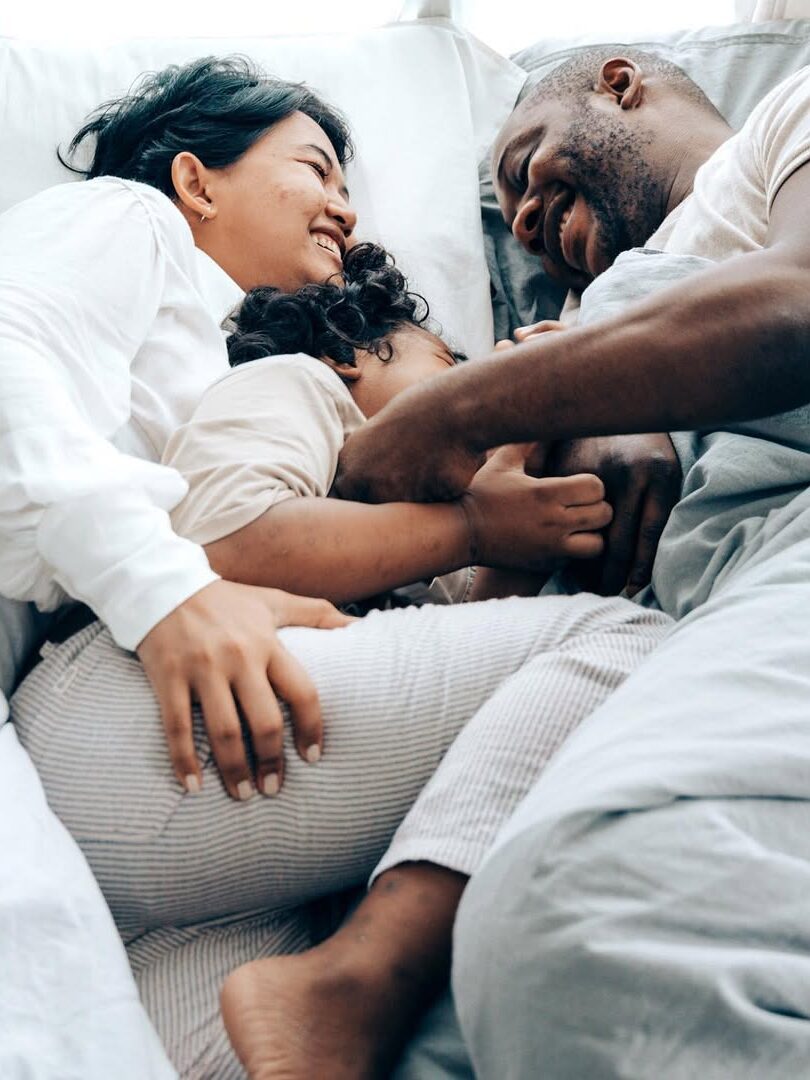 A family of three, dressed in white, cuddle joyfully in bed under a gray blanket, showing smiles and affection.