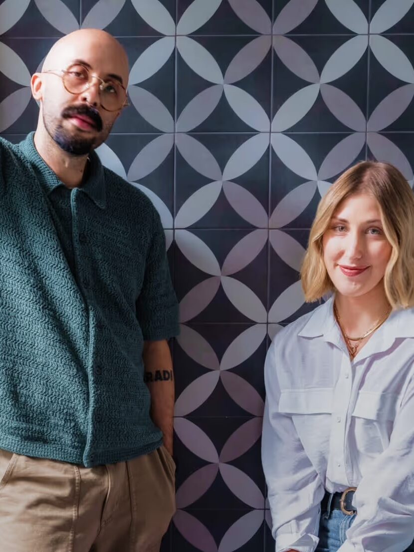 A man in a green shirt and glasses leans against a wall beside a woman in a white blouse. They are in front of a geometric patterned background.