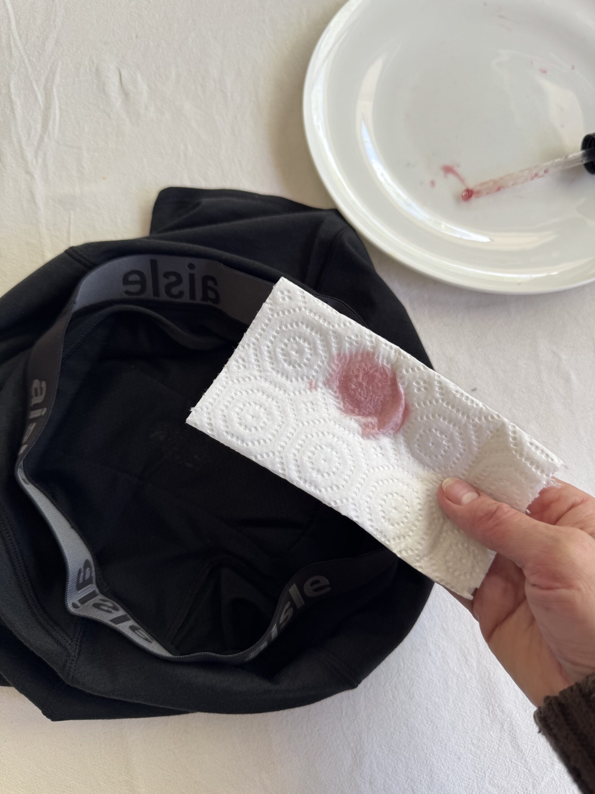 A hand holds a paper towel with a red stain. A black garment and a white plate with a similar stain are visible on a white surface.