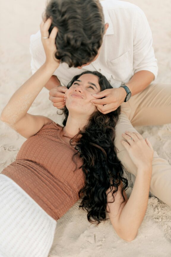 A woman lies on sand with her head in a man's lap while he gently pulls her cheeks. Both appear relaxed and casual.