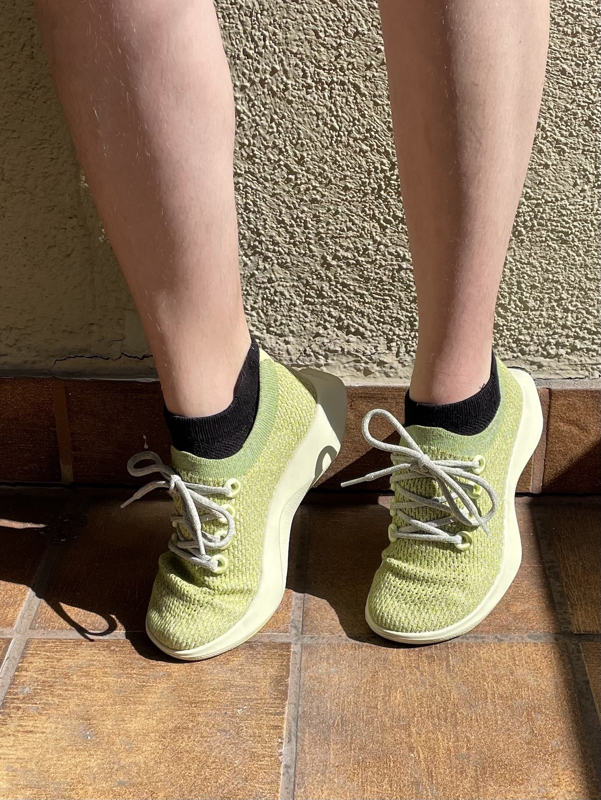 Person standing on brown tile floor wearing light green sustainable sneakers with white laces and black ankle socks, as sunlight casts soft shadows across their legs and shoes.