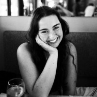A woman with long hair smiles at the camera, resting her head on her hand while sitting at a table in a restaurant. A glass of water is in the foreground.