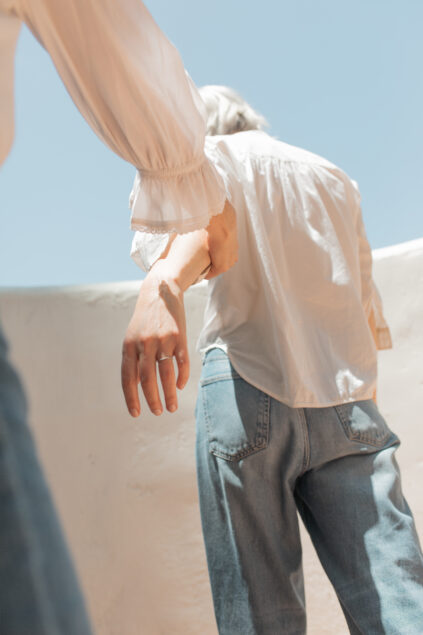 Two people in light clothing and jeans, one holding the other's hand, walk outdoors under a clear sky. The scene is bright and sunlit.