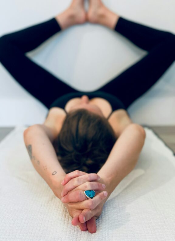 Person lying on their back on a mat with legs spread against a wall and arms extended overhead, hands clasped, wearing black leggings and a blue ring.