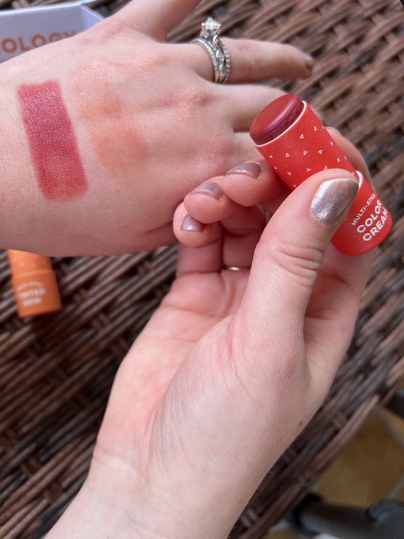 Two hands, one with swatches of makeup on the back, the other holding a small red makeup product tube. The background is a woven surface.