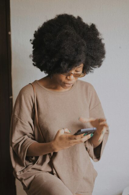 A person with curly hair and glasses is wearing a loose, beige outfit and using a smartphone while standing indoors against a plain, light-colored wall.