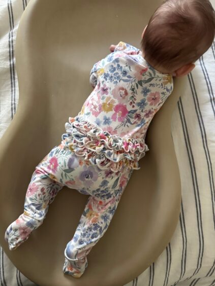 A baby in a floral outfit with ruffles lies face down on a tan changing pad atop a striped fabric surface.