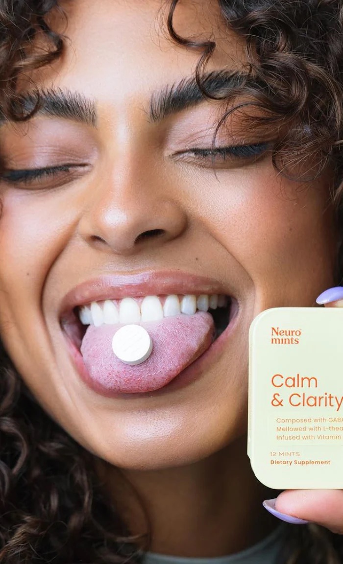 A person with curly hair holds out their tongue with a mint on it and displays a container labeled "Neuro mints Calm & Clarity" in the foreground.
