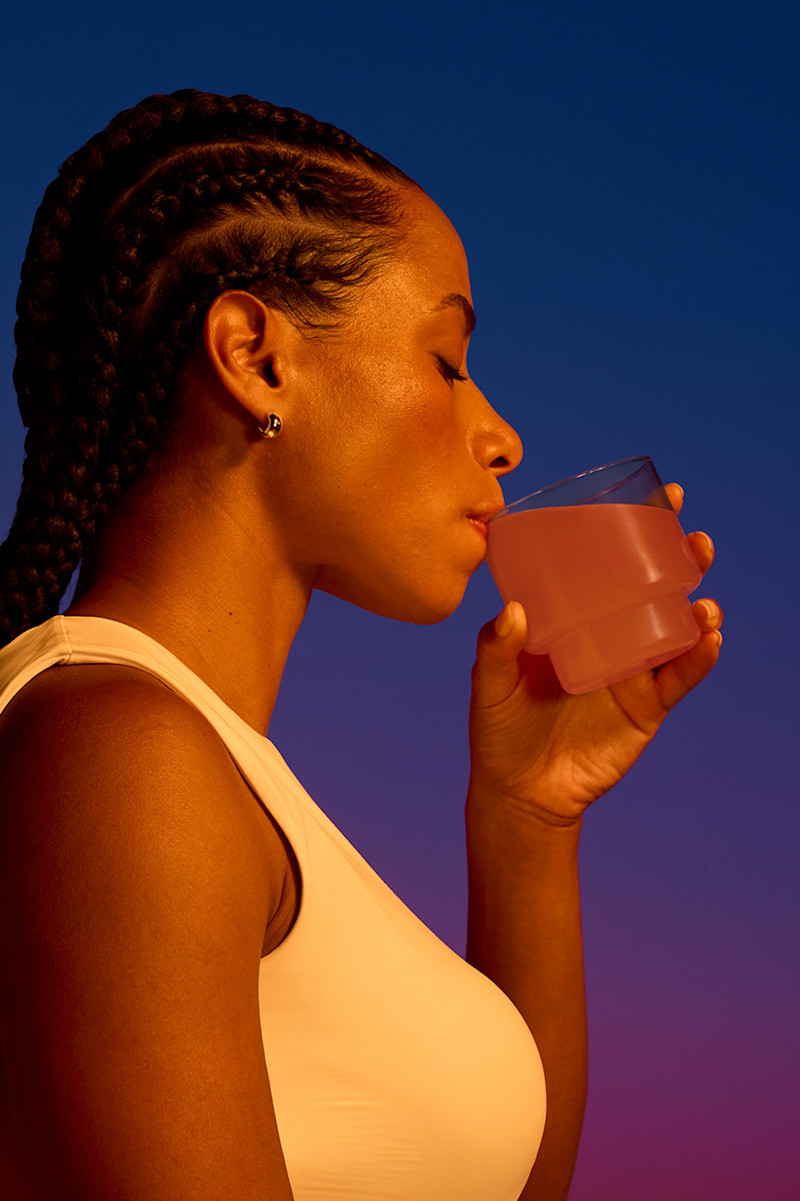 A person with braided hair and a sleeveless top is drinking a pink beverage from a glass against a gradient blue and purple background.
