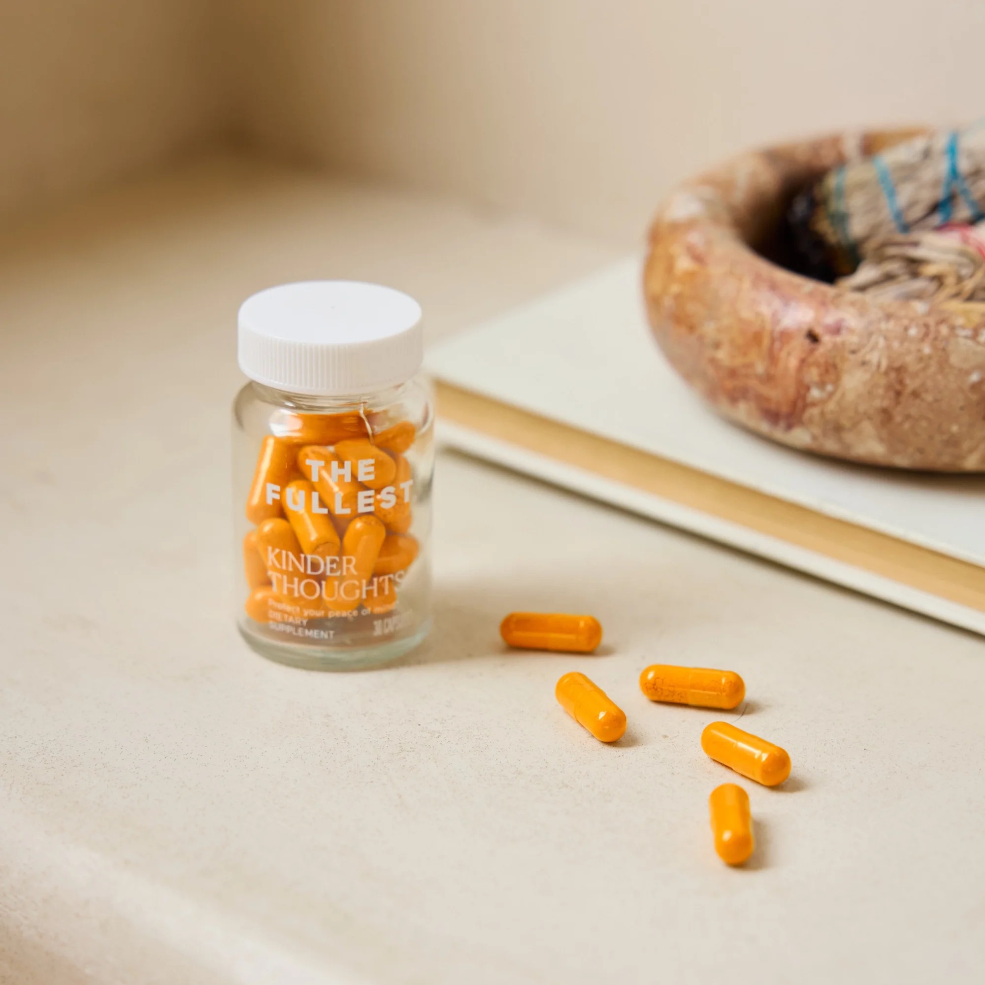 A jar labeled "THE FULLEST KINDER THOUGHTS" dietary supplement sits on a surface with several orange capsules scattered nearby.