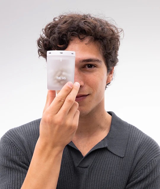 A man with curly hair in a dark shirt holds a THESIS-branded translucent container partially in front of his face against a plain background.