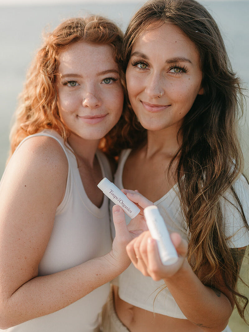 Two women standing by water, holding lip balm tubes labeled "Tropic Therapy," smiling at the camera.