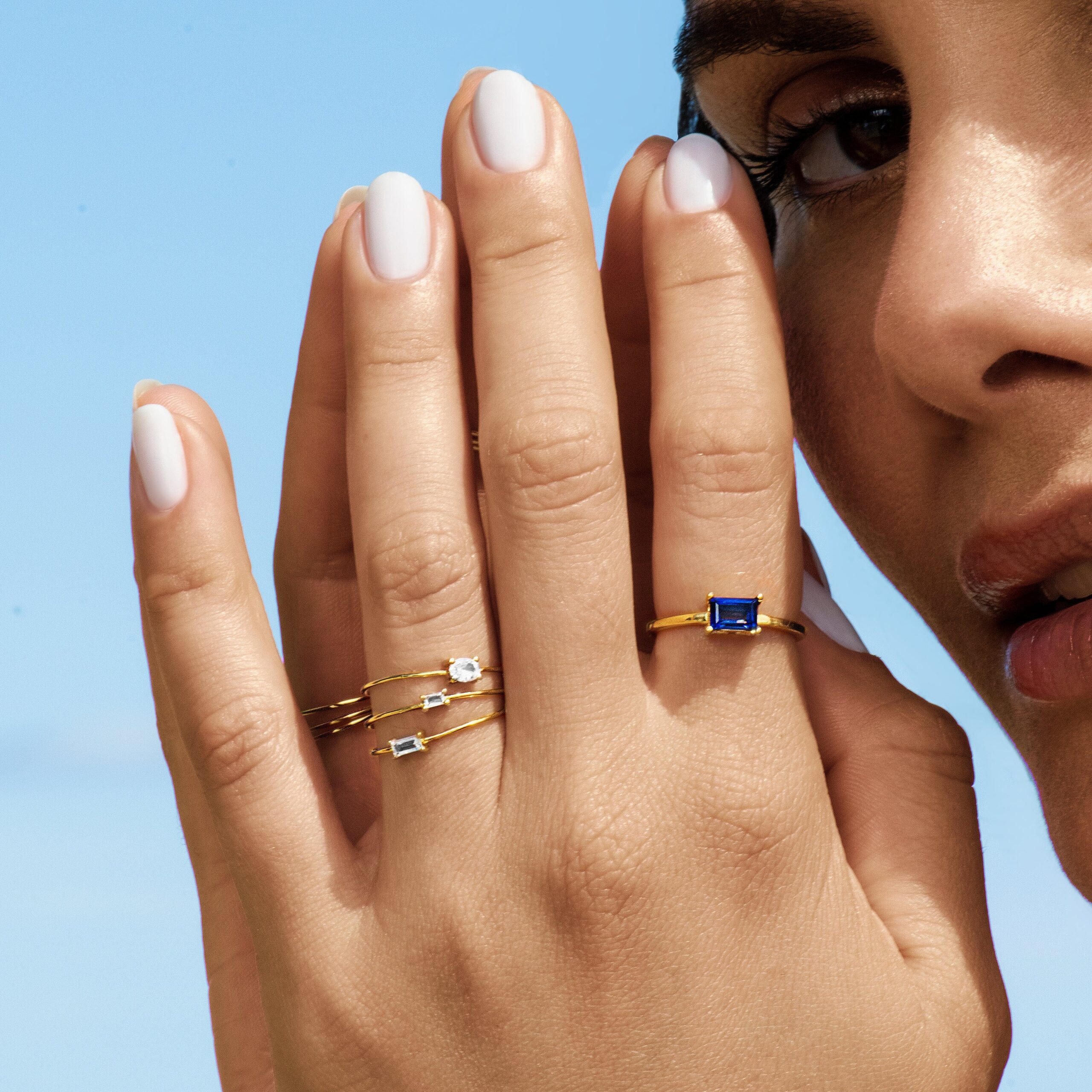 A person with light nail polish wears multiple gold rings with blue and clear stones, holding their hand near their face against a blue sky background.