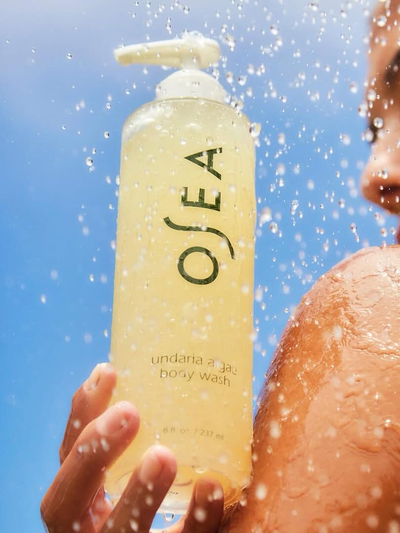 Close-up of a person in the shower holding a bottle of OSEA Undaria Algae Body Wash, with water droplets cascading.
