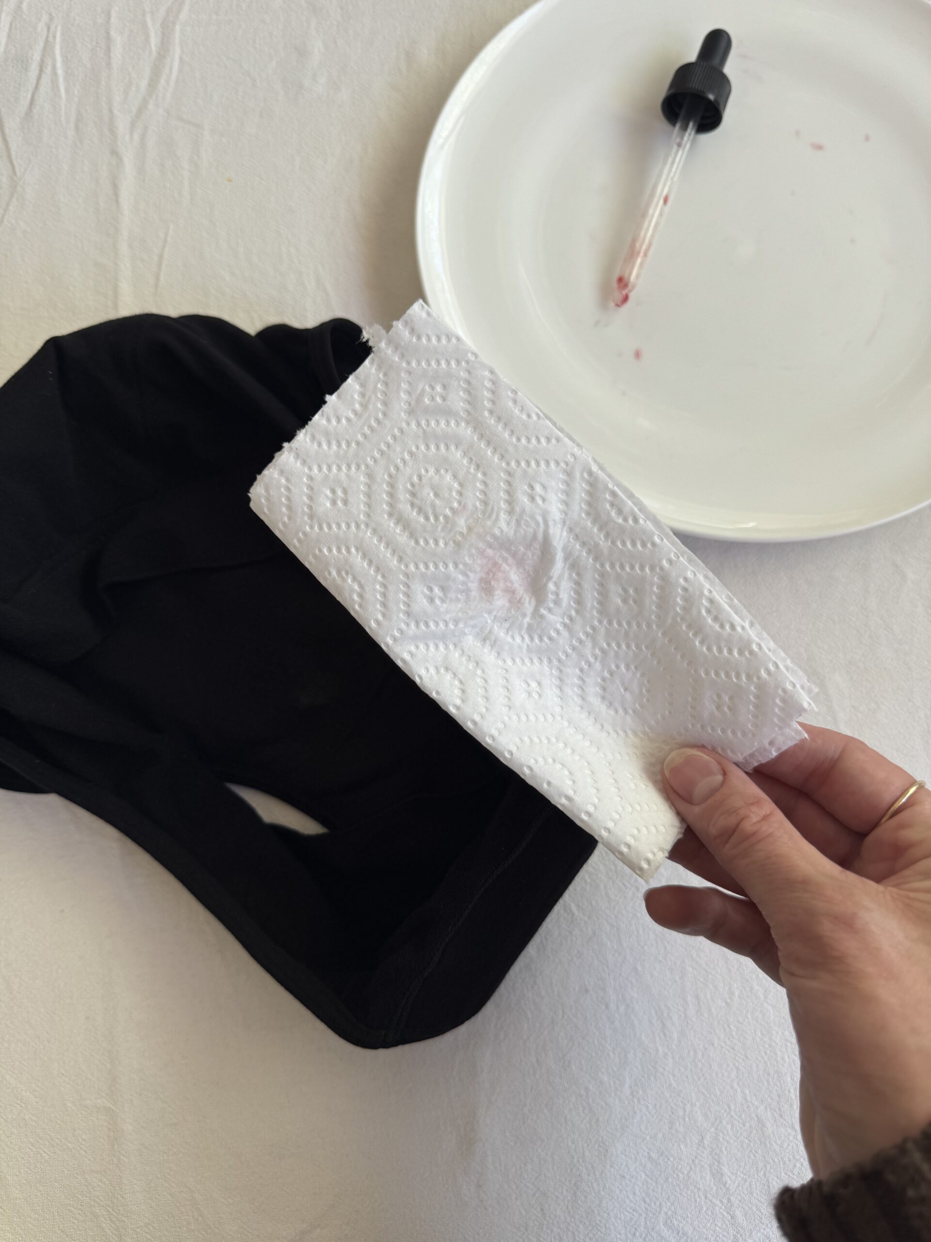 A hand holding a paper towel with a red stain above a black fabric item. Nearby, a white plate with a dropper containing red liquid.