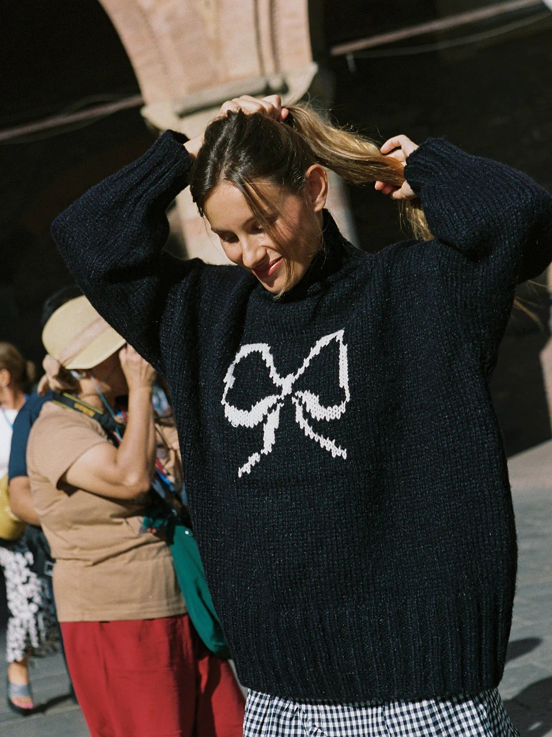 A woman outdoors ties her hair back while wearing a black sweater with a white bow design; other people are visible in the background.