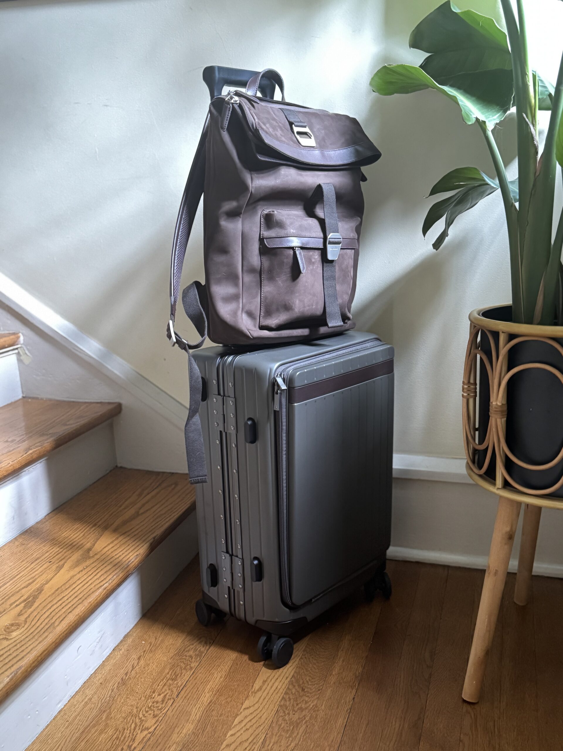 A brown backpack rests on top of a gray hard-shell suitcase with wheels, placed next to a staircase and a potted plant indoors.