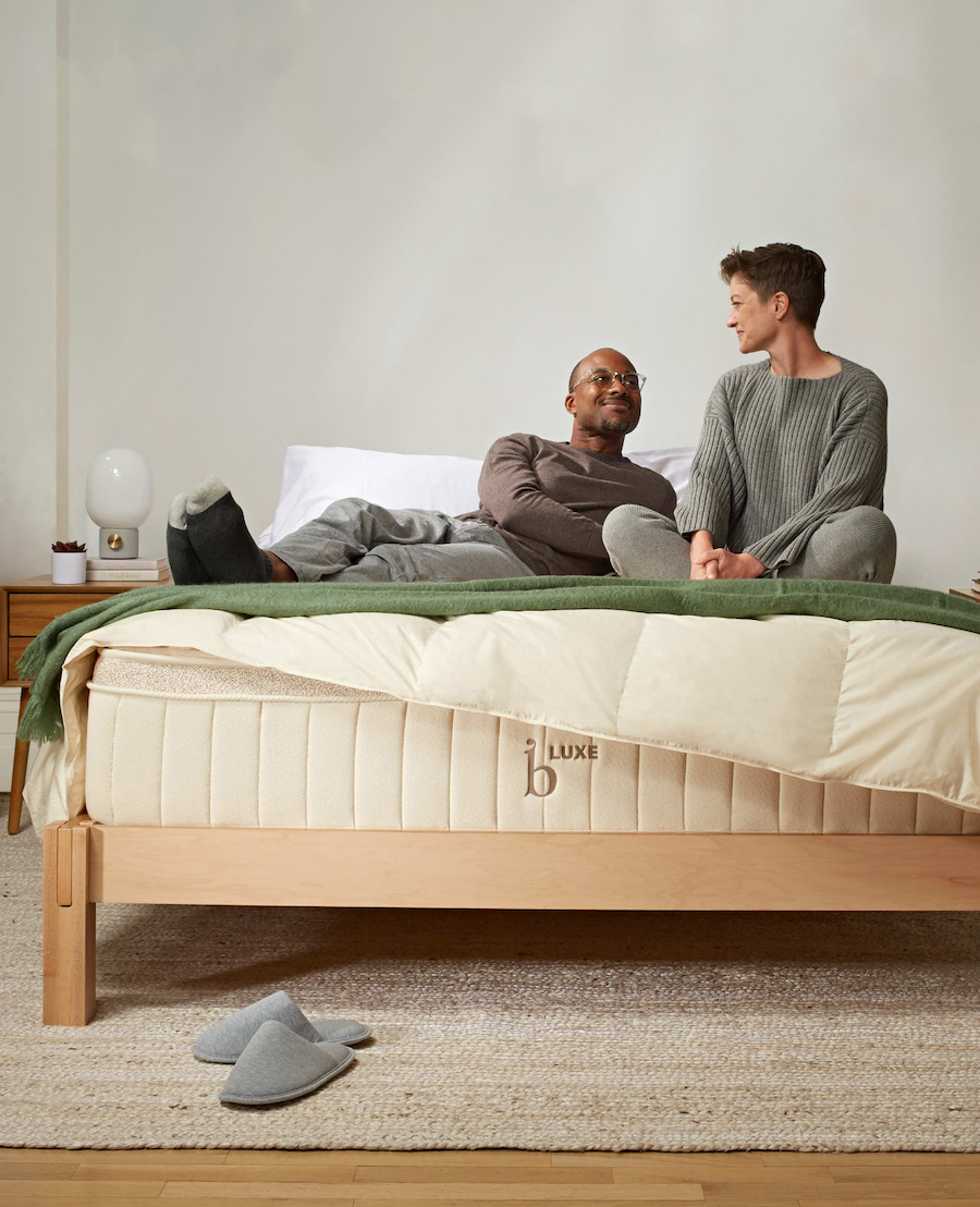Two people in casual clothes relax on a Birch bed with a wooden frame, enjoying the comfort of an organic mattress. Gray slippers are on the floor, and a white nightstand with a lamp is beside the bed.