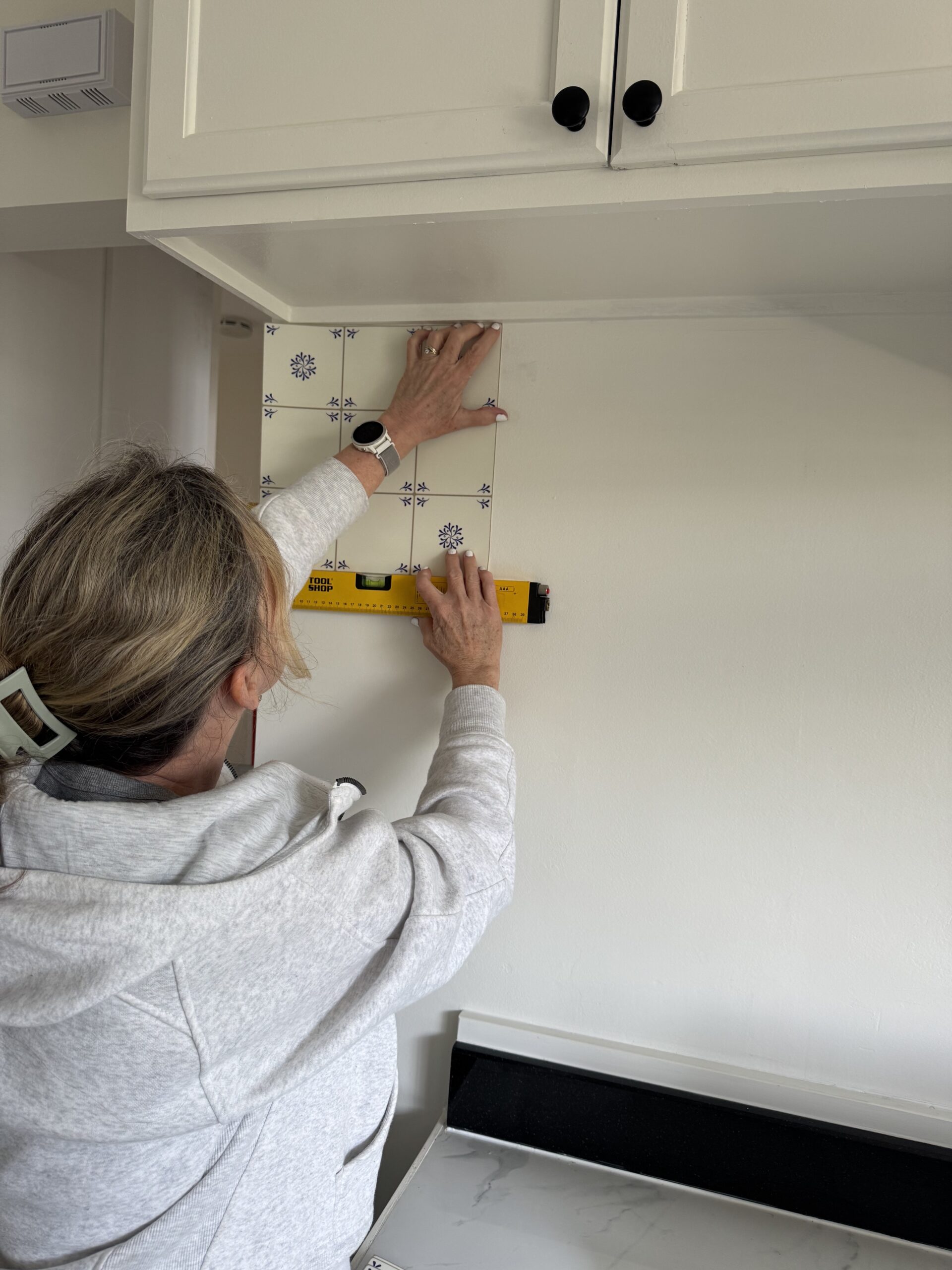 A person measures and aligns decorative tile stickers on a kitchen wall beneath a cabinet using a yellow level tool.