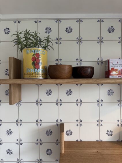 A wooden shelf holds a rosemary plant in a tomato can, two wooden bowls, and a box against a blue and white patterned tile wall.