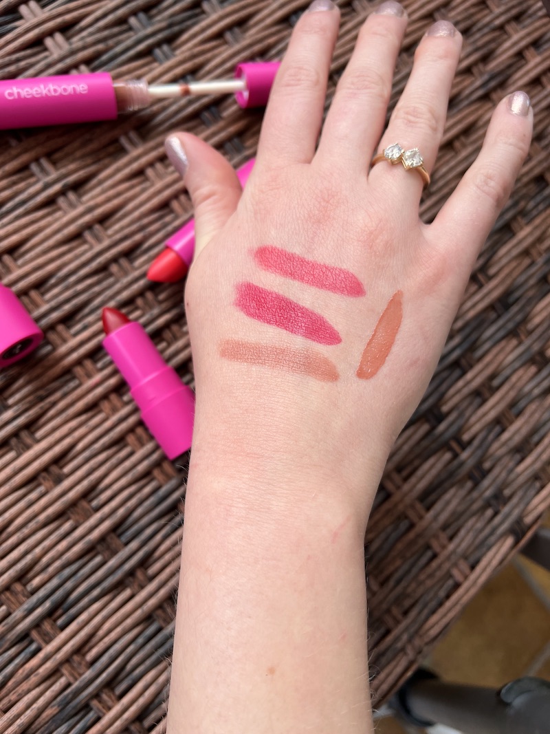 A hand with swatches of four different lip colors on the back, with various pink and red lipsticks shown next to it on a woven surface. The hand has a gold ring with a diamond.
.