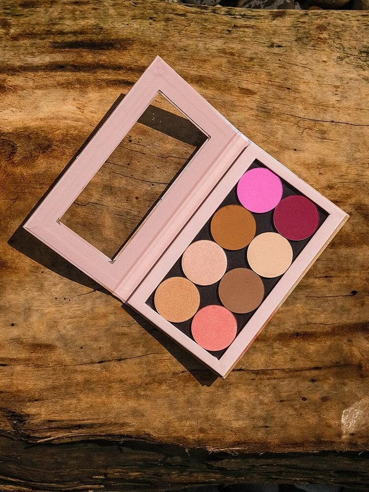 An open eyeshadow palette with nine colors, including various shades of brown, pink, and red, placed on a wooden surface.