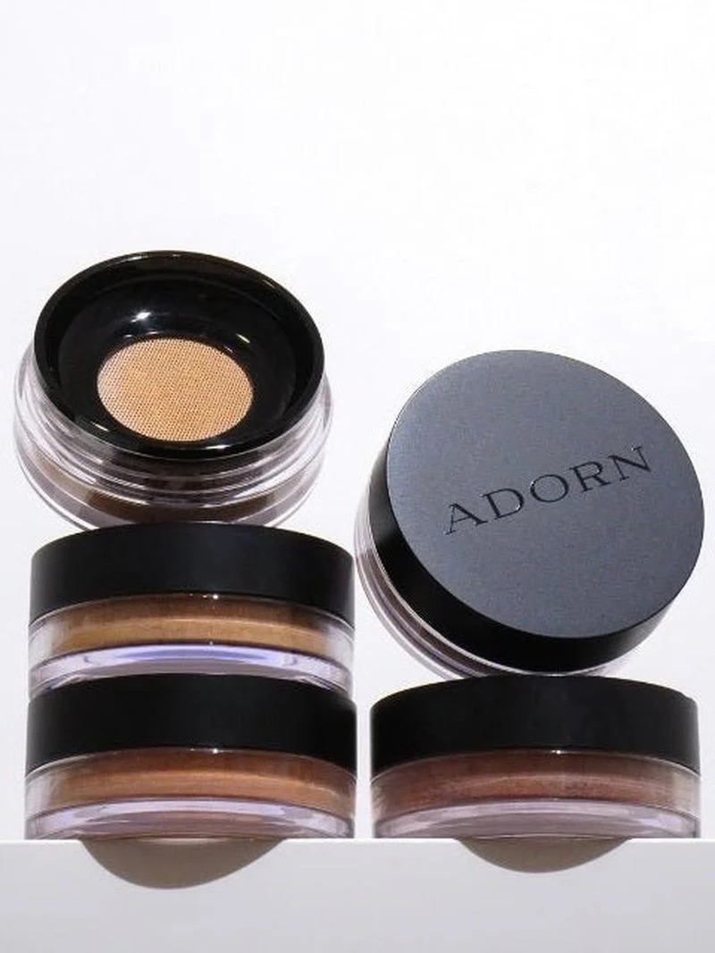 Four small round containers of Adorn mineral makeup are stacked, with one lid off to reveal a pressed powder inside.