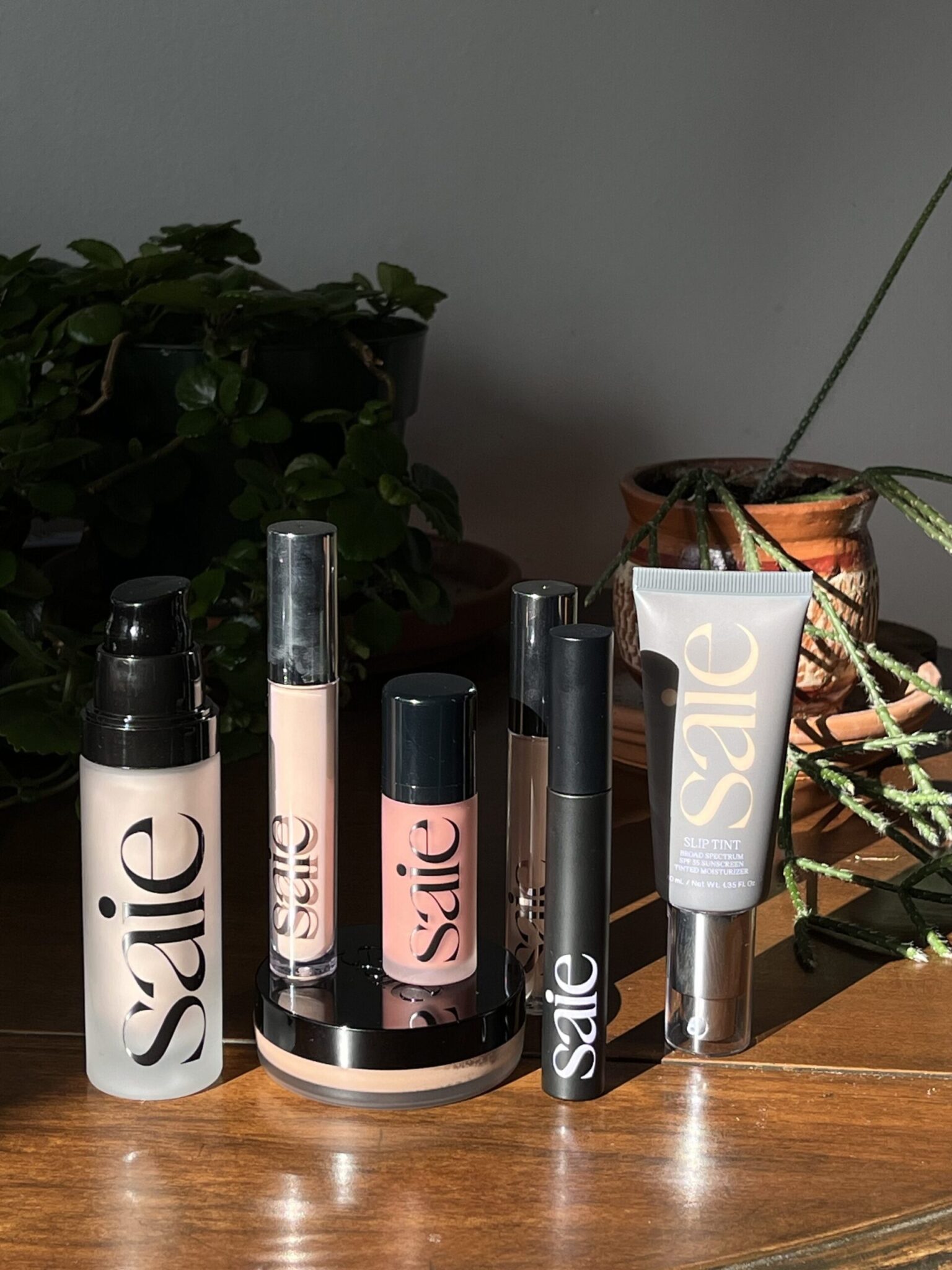 A collection of Saie makeup products, including foundation, concealer, mascara, and lip gloss, arranged on a wooden table with potted plants in the background.