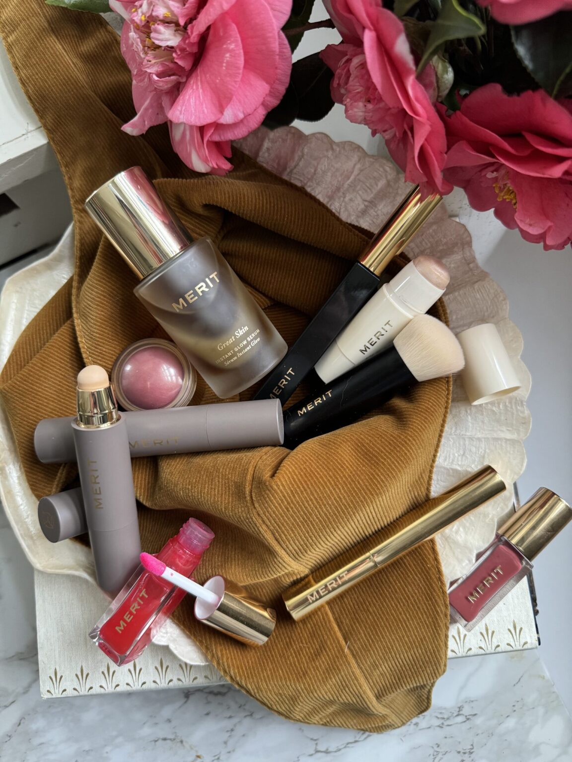 Makeup products arranged on a brown fabric with pink flowers nearby. Includes foundation, blush, lip gloss, and brushes marked "MERIT.
