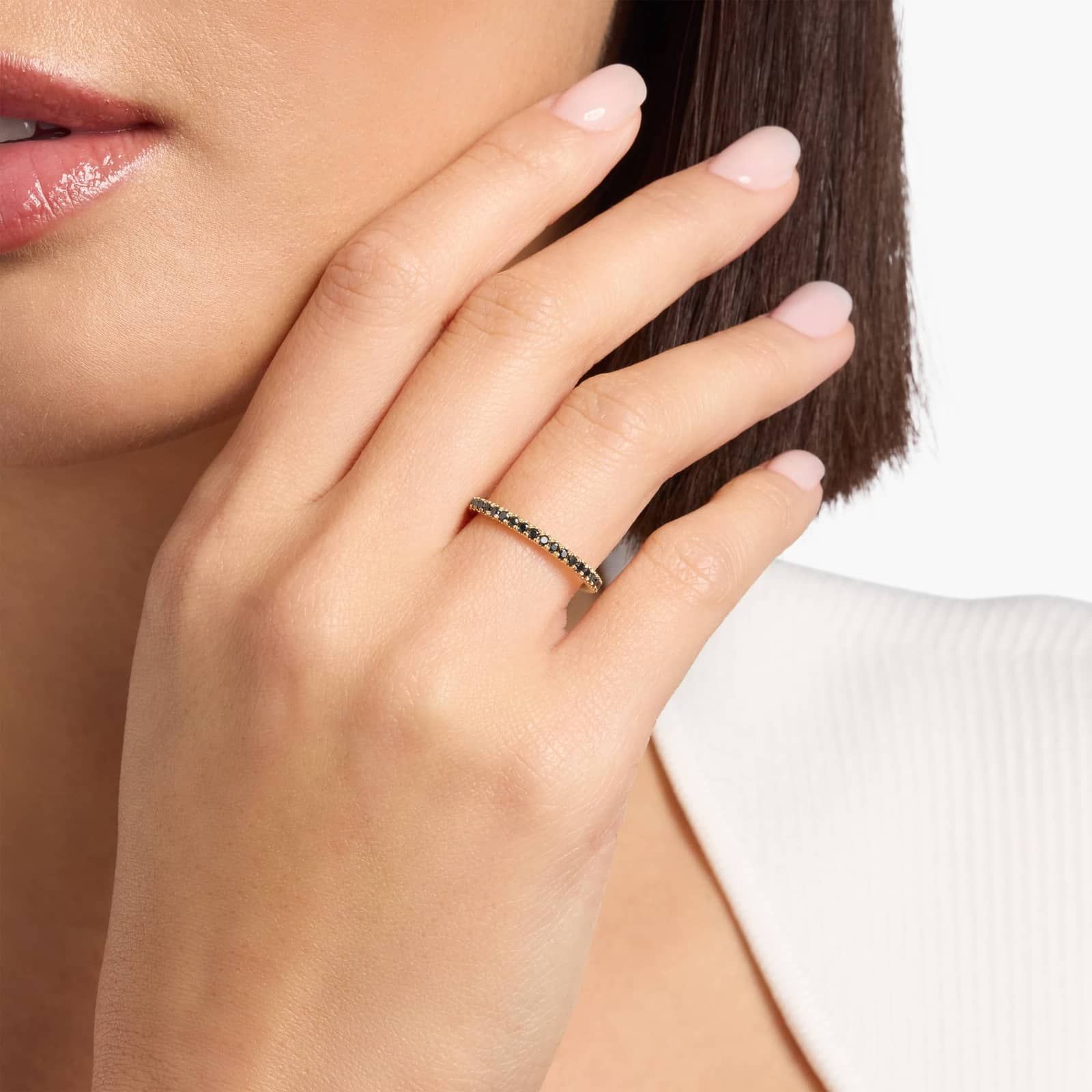 A woman's hand with neatly manicured nails touches her face, showcasing a thin gold ring with small dark stones on her finger.