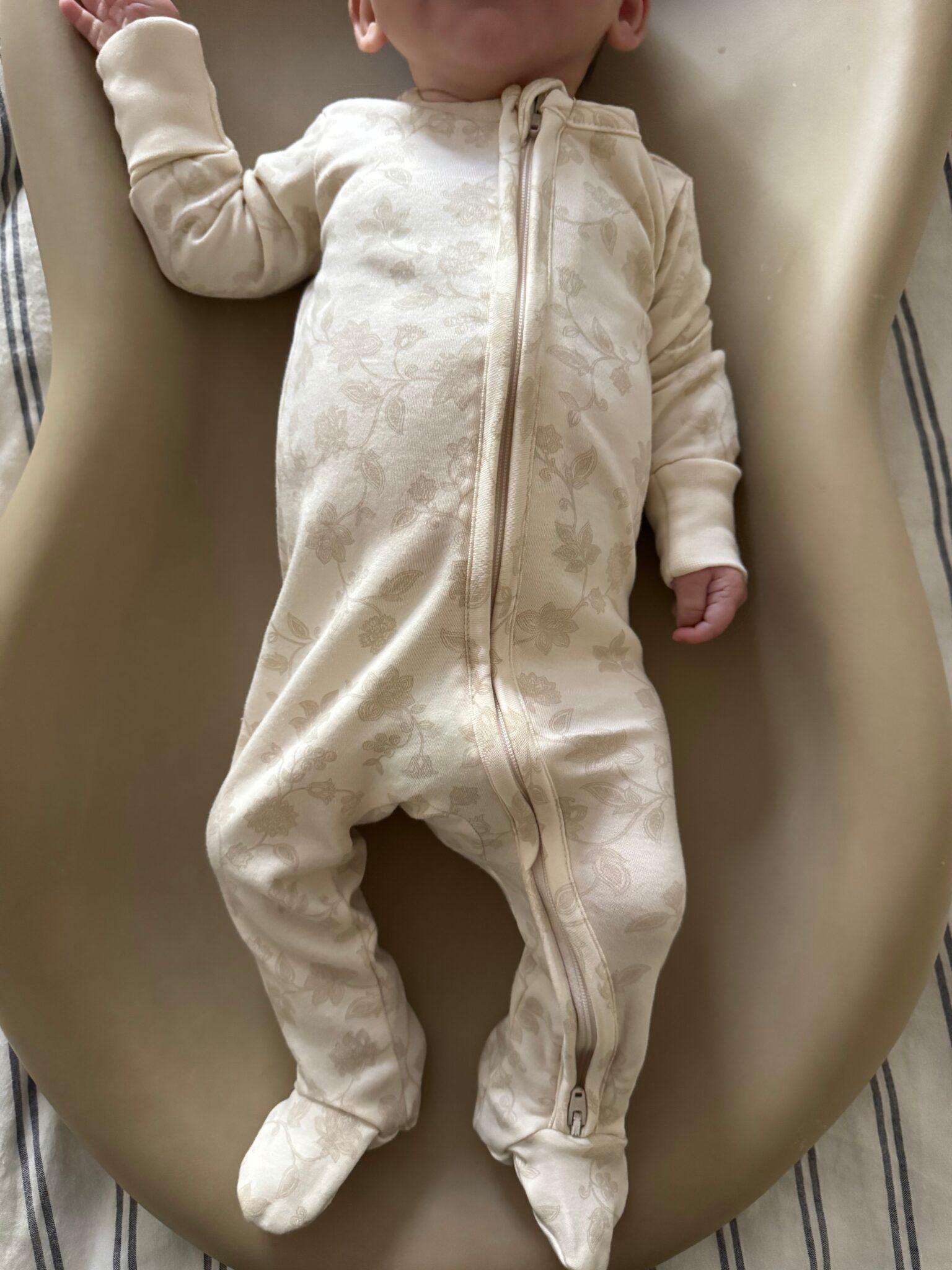 A baby in a cream-colored, floral-patterned zip-up onesie lies on a beige surface, with one arm raised and face partially out of frame.