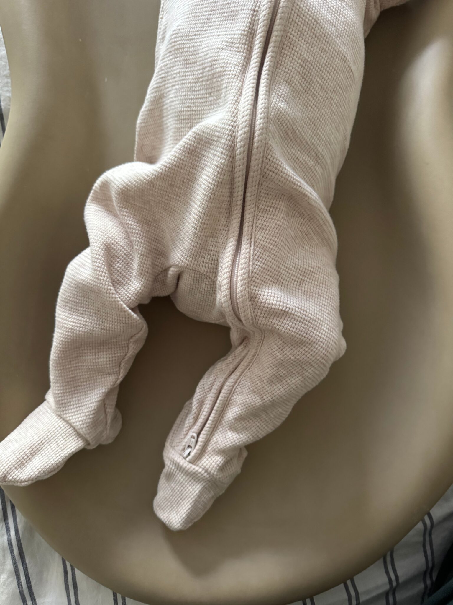 A baby wearing a light textured onesie lies face down on a taupe-colored surface, with only the lower half of the body visible.