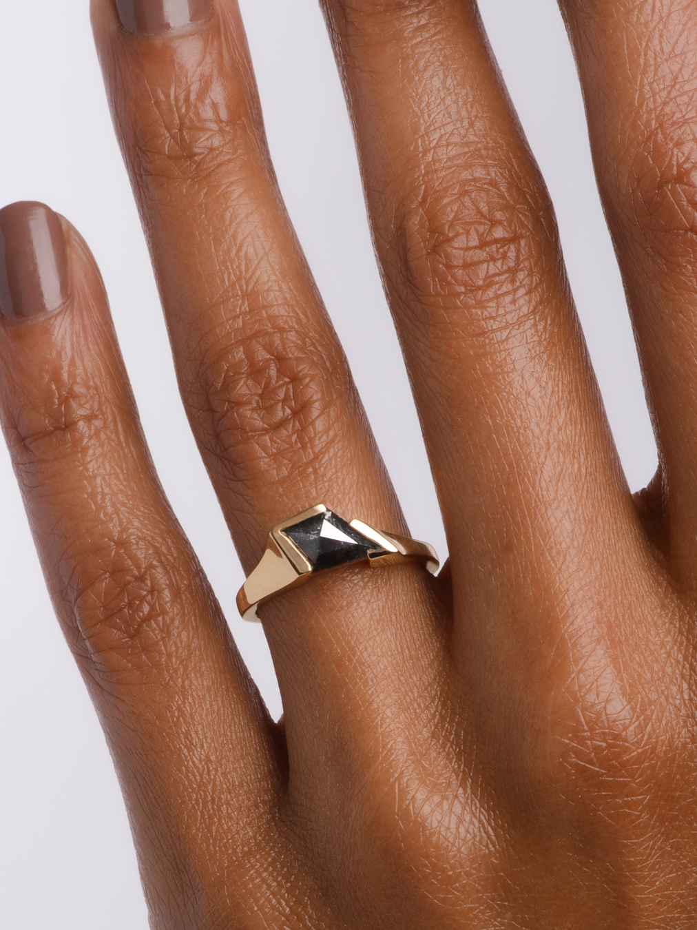 A hand with neutral nail polish wears a gold ring featuring a geometric black stone on the ring finger.