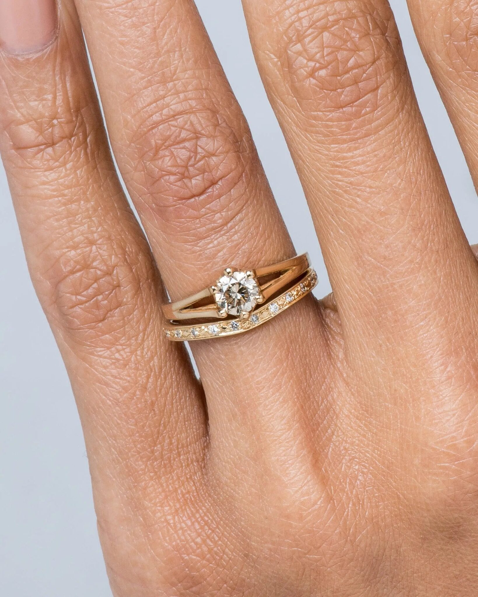 A hand wearing two stacked gold rings, one with a central round diamond and one with small diamonds set along the band.