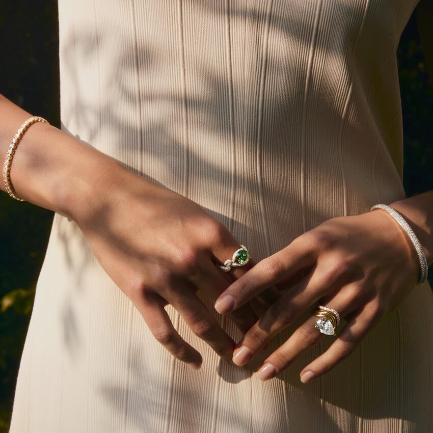 A person in a beige, ribbed dress displays multiple rings and bracelets on both hands, with sunlight casting soft shadows over their arms.