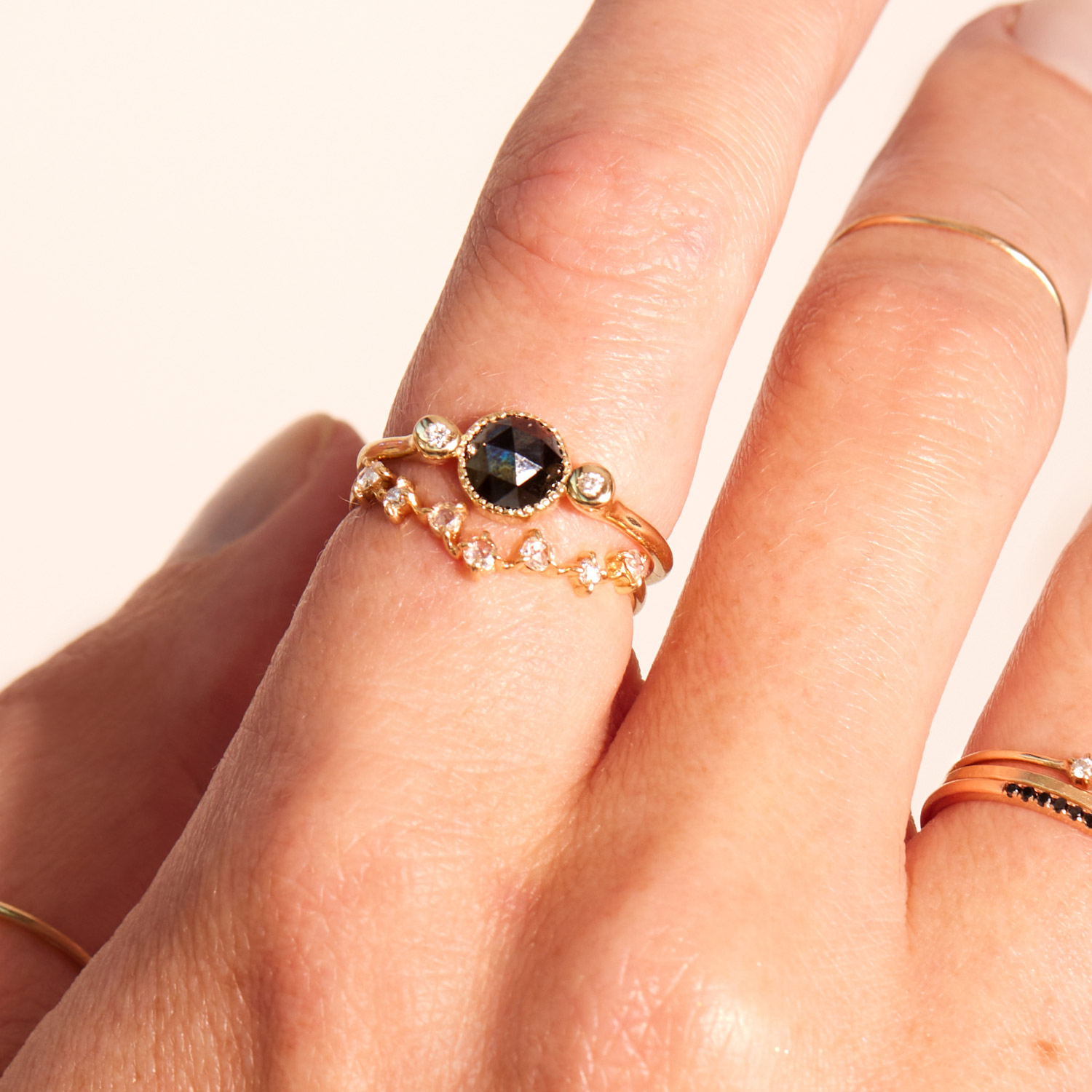 A hand wearing gold rings, one with a large black gemstone and small diamonds, and another thin gold band with small clear stones in a curved shape.