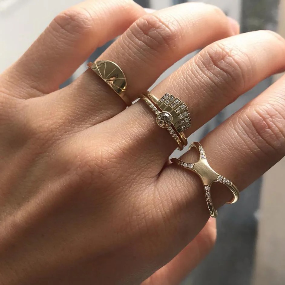 A hand wearing four gold rings, each with unique designs, including geometric shapes and small embedded diamonds, on the index, middle, and ring fingers.