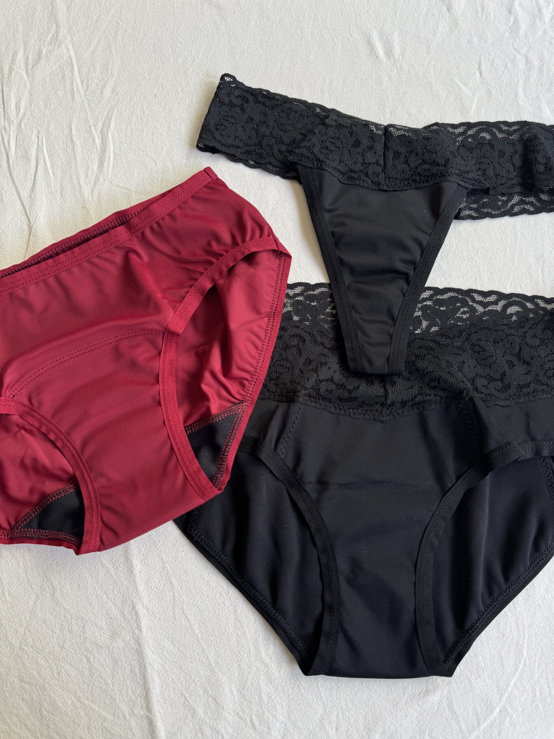 Three pairs of women's underwear: one red pair and two black pairs with lace, arranged on a white fabric surface.
