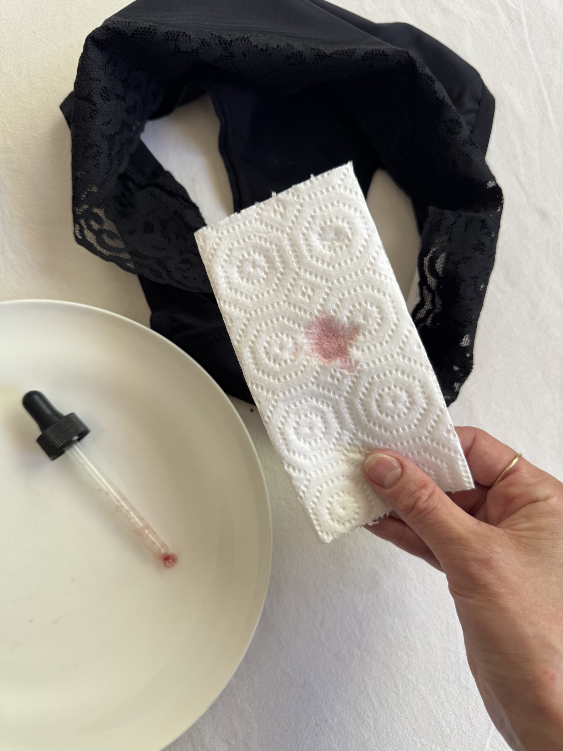 Hand holding a paper towel with a red stain over a plate containing a dropper, next to black lace fabric.